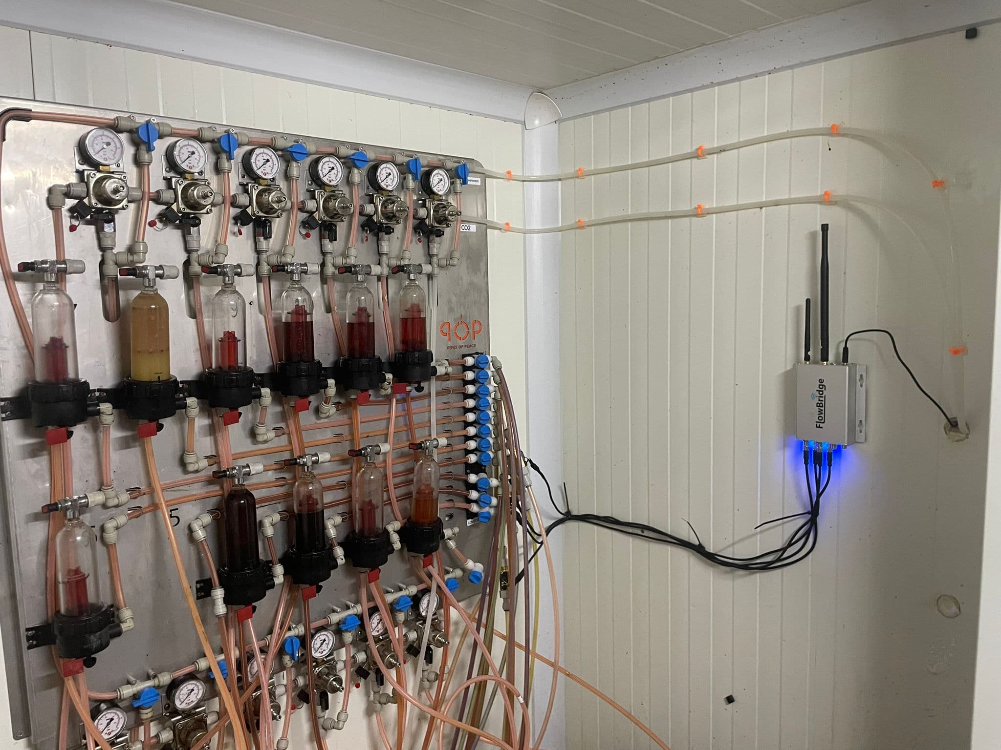 A FlowBridge gateway mounted on a cellar wall, indicator lights lit, cabling running across to the beer lines and gas manifold